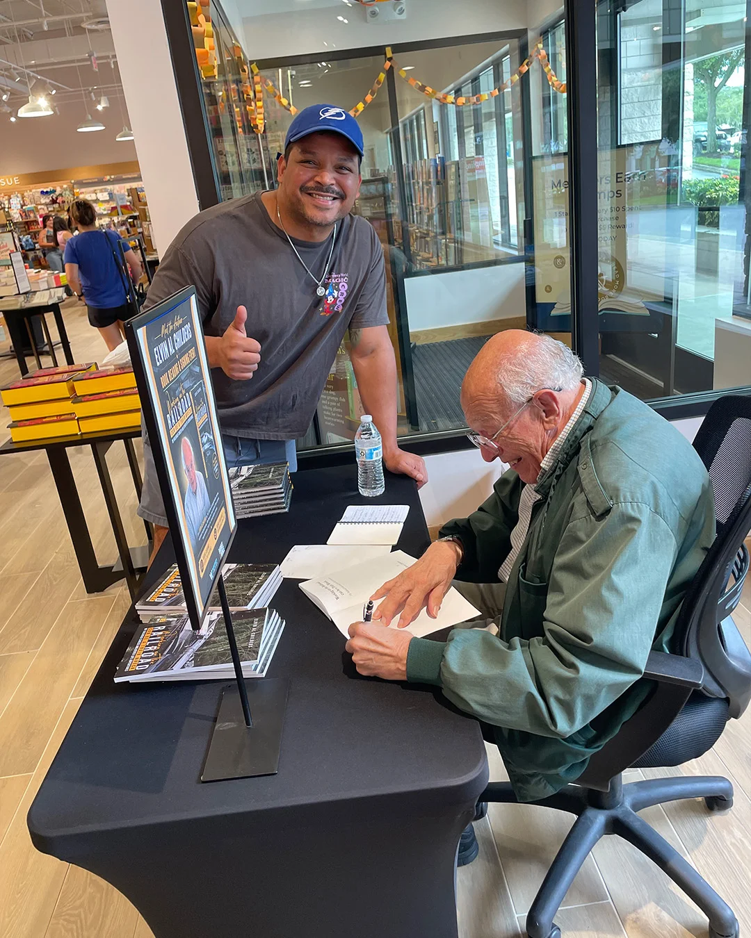 A reader giving a thumbs up across the table as the author signs a copy for him