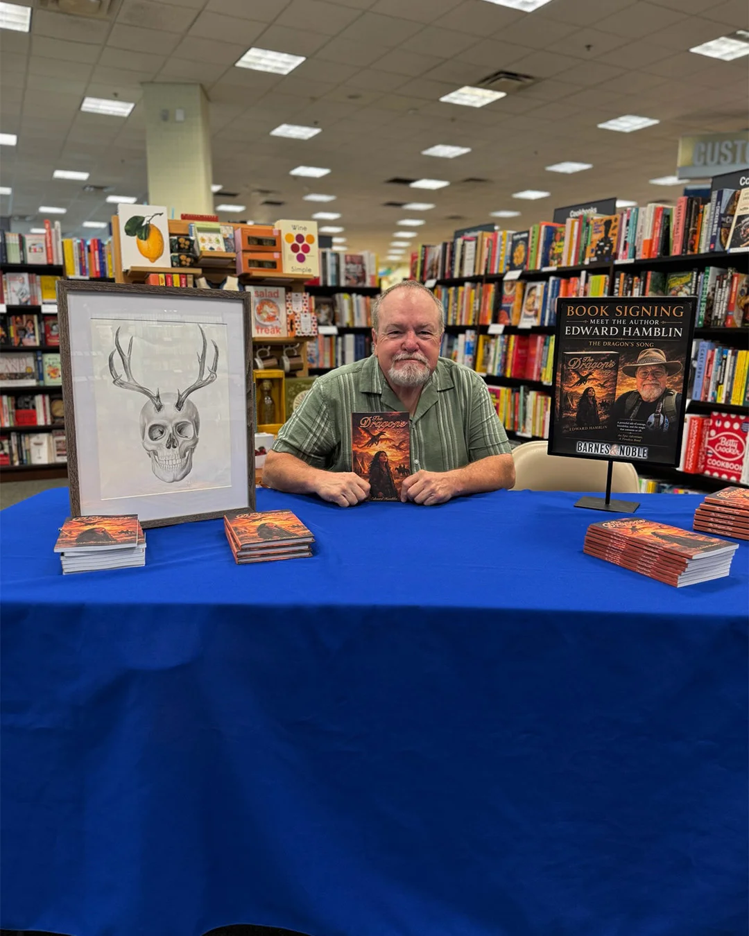 An author holding up his book behind a signing table set with stacked copies, framed cover artwork and a book signing poster